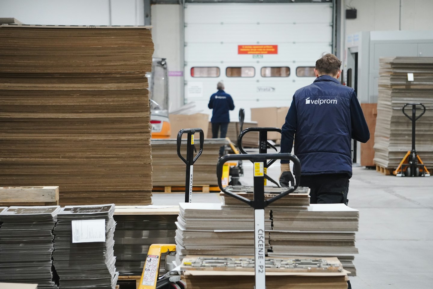Men operate pallet jacks in a busy warehouse with stacked cardboard.